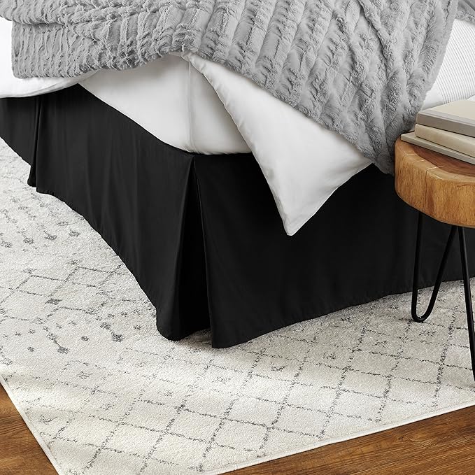 Bed with black skirt, gray textured blanket, and wooden side table on a patterned rug.