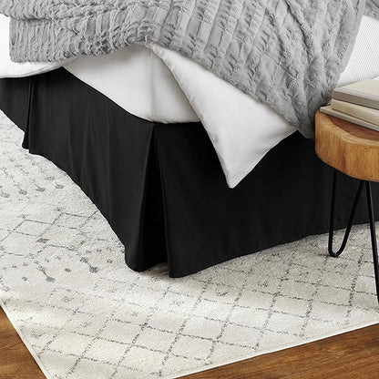 Bed with black skirt, gray textured blanket, and wooden side table on a patterned rug.