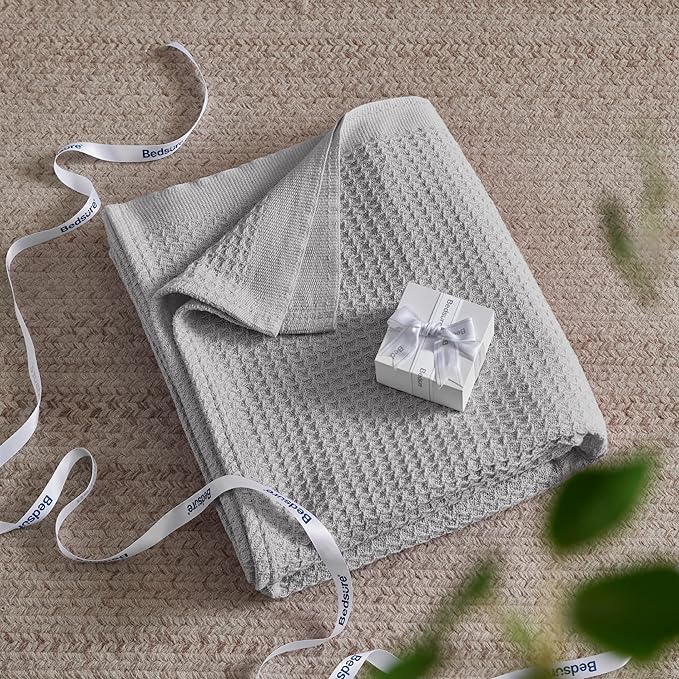 Gray textured blanket with a small white gift box on a beige surface, featuring a ribbon.